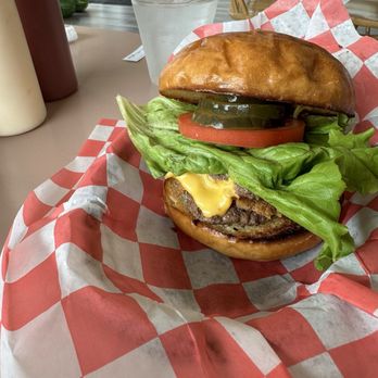 White Lightning Restaurant - Smurd Burger - A cheeseburger with lettuce, tomato, pickles, and melted cheese on a toasted bun, placed on red and white checkered paper, with a glass of water and condiments in the background. - American Near Me - Lampe, Missouri