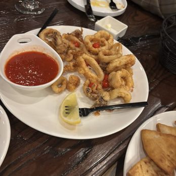 The Foundry - Calamari - A white round plate holds fried calamari rings garnished with small cherry tomatoes. A small bowl of red dipping sauce is placed on the plate, along with a lemon wedge. The plate is on a dark wooden table, with a person in a plaid shirt using utensils in the background. Additional dishes and glasses are visible on the table, indicating a dining setting. - New American Near Me - Hicksville, New York