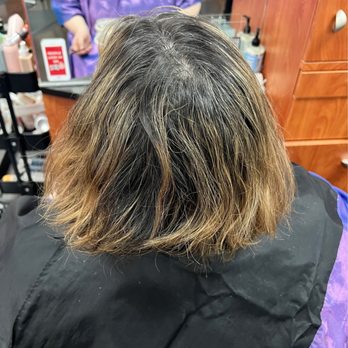SYS Hair - Before - The image shows the back of a person's head with shoulder-length, wavy hair. The hair has dark roots transitioning into lighter, blonde highlights. The person is seated in a salon chair, with salon equipment and a stylist visible in the background. The setting appears to be a hair salon, with wooden cabinets and hair products nearby. - Hair Salons Near Me - Phoenix, Arizona
