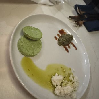 Joon - The image shows a white oval plate containing three distinct food items: two green hummus portions, a meatball topped with a drizzle of sauce, and a white cheese dish garnished with herbs and olive oil. The plate is placed on a table with a white tablecloth, alongside utensils, napkins, and glasses, indicating a dining setting. - Persian/Iranian Near Me - Vienna, Virginia