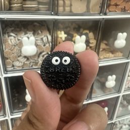 A black beaded brooch featuring a face with white beaded eyes and a textured surface is held between fingers. The background shows a display case with multiple small decorative items, including bunny-shaped figures and other small objects, arranged in compartments. The image emphasizes the brooch's detailed beadwork and face design.