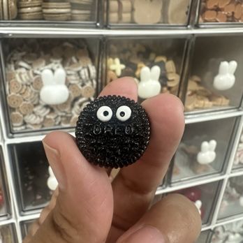 Lumee - A black beaded brooch featuring a face with white beaded eyes and a textured surface is held between fingers. The background shows a display case with multiple small decorative items, including bunny-shaped figures and other small objects, arranged in compartments. The image emphasizes the brooch's detailed beadwork and face design. - Arts & Crafts Near Me - Pasadena, California