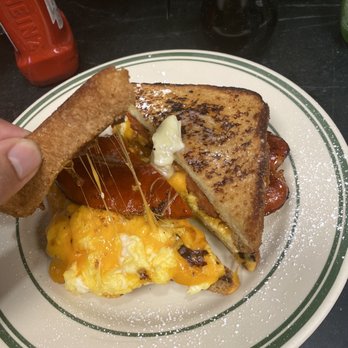 Ray's Luncheonette - French toast sandwich - A breakfast plate contains scrambled eggs with melted cheese, crispy bacon, and a grilled sandwich with toasted bread. A hand is holding a piece of toast above the plate, with melted cheese stretching from the toast to the eggs. The plate is on a dark surface, with a ketchup bottle and a coffee pot visible in the background. - Breakfast & Brunch Near Me - Montclair, New Jersey