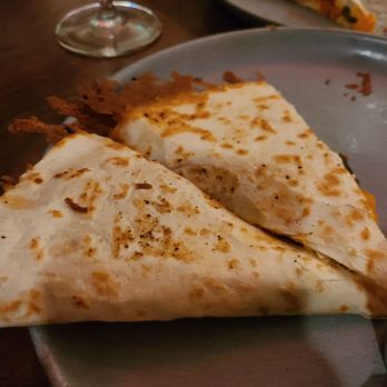 Para Maria - Shrimp quesadilla - Two slices of quesadilla with melted cheese and a slightly browned, crispy exterior are served on a gray plate. A glass of wine is partially visible in the background, along with another slice of quesadilla on a separate plate. The setting appears to be a dining table, likely in a restaurant or home environment. - Wine Bars Near Me - Boston, Massachusetts