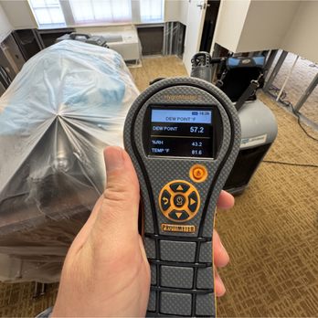 Quality Mold Solutions - A hand holds a digital hygrometer displaying dew point, humidity, and temperature. The background shows a room with covered f... - Environmental Testing Near Me - Orlando, Florida Quality Mold Solutions - A hand holds a digital hygrometer displaying dew point, humidity, and temperature. The background shows a room with covered f... - Environmental Testing Near Me - Orlando, Florida