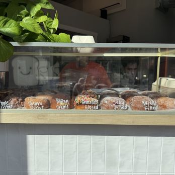 Moore Bread - Jelly Donut (GF) Cinnamon Roll (GF) Boston Creme (GF) - A glass display case contains gluten-free donuts labeled with handwritten tags, including flavors like Boston creme and jelly filled, with a plant and some interior elements visible. - Bakeries Near Me - Denver, Colorado