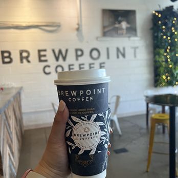 Brewpoint Craft Elmhurst - A hand holding a Brewpoint Coffee cup is shown inside the cafe, with a wall sign reading 'BREWPOINT COFFEE' in the background. - Coffee & Tea Near Me - Elmhurst, Illinois
