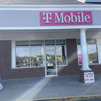 T-Mobile - Outside - The image shows the exterior of a T-Mobile store with a pink sign displaying the company logo and name. The storefront features large glass windows and a glass door, with a brick facade and a white overhang supported by a column. There is a sidewalk and parking lot in front of the store, with a sign indicating parking restrictions. The store is situated in a commercial shopping area. - Mobile Phones Near Me - Cranston, Rhode Island