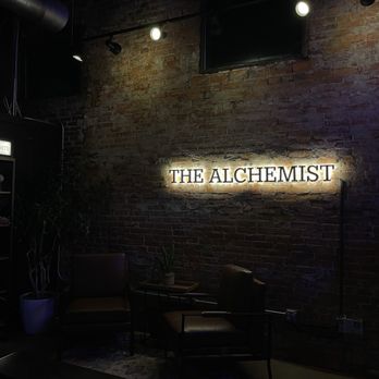 The Alchemist - A seating corner with two chairs and a small table, set against a brick wall with a backlit sign that says 'The Alchemist' in Indianapolis, IN. - Coffee & Tea Near Me - Indianapolis, Indiana