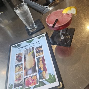 Sugarcane - A bar counter features a pink cocktail in a coupe glass garnished with a lemon wedge and two black straws. Next to it is a tall glass of water with ice and a straw, placed on a black napkin. A menu with tropical-themed images and the text 'Sugarcane BK' is also visible on the counter. - Caribbean Near Me - Brooklyn, New York