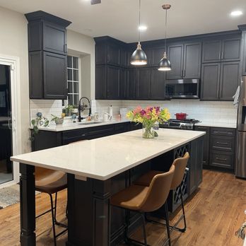 Cabinet Tree Studio - The kitchen has dark gray or black cabinetry with multiple upper and lower cabinets, a white countertop, and a white subway tile backsplash. A large white island with seating for three is centered in the space, with a vase of flowers on top. Pendant lights hang above the island, and a ceiling fan is installed on the ceiling. The kitchen includes a stainless steel refrigerator, microwave, and a window above the sink with plants on the windowsill. The flooring is wood, and the overall design is modern and functional. - Interior Design Near Me - Plainfield, Illinois