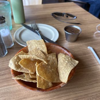Los Arcos - Small chips and salsa. $3 - A wooden bowl containing tortilla chips is placed on a restaurant table with various condiments and utensils. - Tacos Near Me - Chicago, Illinois