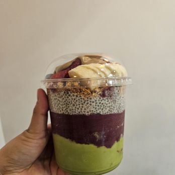 Roots & Rye - Base: Matcha, Acai, and Chia Pudding (ask to sample the bases to see what you like!) - A multi-layered acai bowl in a clear plastic cup, topped with banana, berries, and other toppings, held by a person's hand. - Acai Bowls Near Me - Pasadena, California