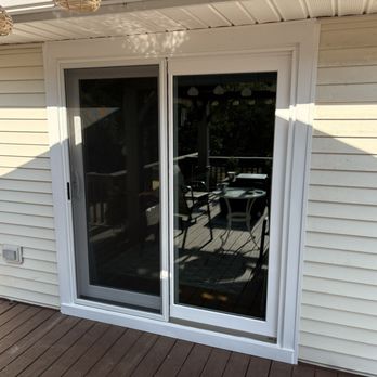 Winchoice - New sliding patio door - A white framed sliding glass patio door is set into the exterior wall of a house, leading to an outdoor deck. The deck has wooden flooring and features a table with chairs, visible through the glass. The house exterior has horizontal siding, and sunlight casts shadows on the wall and deck. - Windows Installation Near Me - Mountain Pine, Arkansas