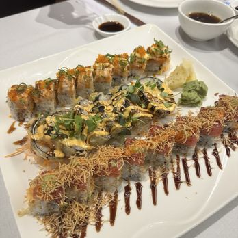 All Seasons Table - A large white square plate holds multiple sushi rolls with various toppings, sauces, and garnishes. The sushi includes rolls topped with crispy flakes, drizzled sauces, and microgreens. Accompanying the sushi are small portions of wasabi and pickled ginger. The setting appears to be a restaurant with a white tablecloth, and in the background, there are bowls of soy sauce, a glass of water, and a person using chopsticks. - Chinese Near Me - Malden, Massachusetts
