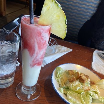 Red Lobster - Sunset Passion Colada - A tall, layered cocktail with a pink and white swirl is on a wooden restaurant table, garnished with a slice of pineapple. The drink has a black straw and is served in a highball glass. Nearby, there is a glass of water and a small salad with lettuce and croutons on a white plate with a blue rim. The background shows part of the restaurant interior with a person and patterned seating. - Seafood Near Me - Albuquerque, New Mexico
