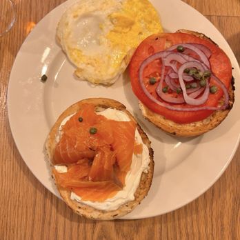 The Foundry Rose - Gravlax Platter - A round white plate holds two bagel toasts topped with smoked salmon, cream cheese, tomato slices, onion rings, and capers. Additionally, there are two fried eggs with slightly runny yolks and cooked whites. The plate is placed on a wooden table, with a glass of water partially visible at the top left corner. - Cafes Near Me - Cold Spring, New York