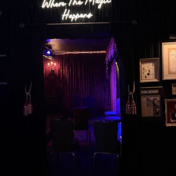 Trickery - An intimate performance space features a small stage with velvet curtains and a neon sign overhead that reads 'Where The Magic Happens.' The stage is lit with colored lighting, and there are chairs arranged in front for viewers. The venue has dark walls decorated with framed pictures and certificates, creating a theatrical atmosphere suitable for a magic or comedy show. - Performing Arts Near Me - Chicago, Illinois