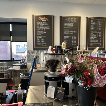 Grinders Coffee House - The image shows the interior of a coffee shop with a counter area. Behind the counter, there are three black menu boards displaying drink options, including coffee, energy drinks, and baked goods. Coffee equipment, such as a grinder and espresso machine, is visible on the counter. A large bouquet of flowers in a black vase is placed on the counter, adding decoration. A window with a partially closed blind is on the left side, allowing some natural light into the space. Various items, including a hand sanitizer bottle and a small sign, are also on the counter. - Coffee & Tea Near Me - Orem, Utah