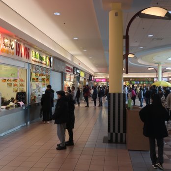 First Markham Place - View of a busy and vibrant food court. - An indoor food court in a shopping mall features several restaurant counters with illuminated signs, including Asian cuisine and other options. Shoppers are walking, standing, and waiting in line, with some seated at tables under yellow umbrellas. The area has tiled floors, yellow columns, and a ceiling with recessed lighting and decorative circular features. The scene captures a typical bustling mall food court environment. - Shopping Centers Near Me - Markham, Ontario