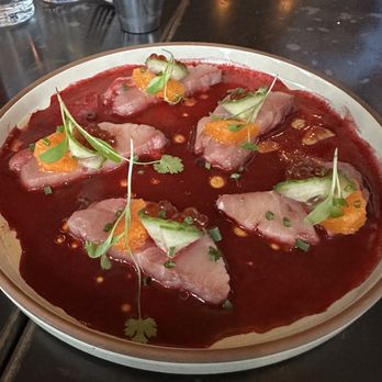 Beach Road - Hamachi Crudo - A plate features hamachi crudo, with thin slices of raw fish arranged on a white dish. The fish is topped with microgreens, orange segments, and cucumber slices, all placed over a red sauce or marinade. The presentation includes small drops of oil or sauce and finely chopped herbs, creating a colorful and detailed seafood dish. - Seafood Near Me - Vineyard Haven, Massachusetts