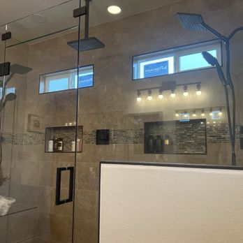 Carpeteria - A bathroom features a glass shower with two showerheads, built-in shelves, and beige tiled walls, with small windows and ceiling lights. - Carpeting Near Me - Lancaster, California