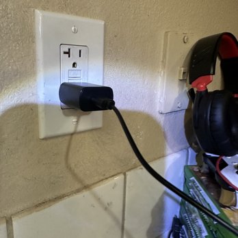  Redd’s Electrical Services - A black electrical device is plugged into a standard wall outlet on a beige wall. The device's cord extends downward, and the shadow of the device is visible on the wall. To the right, there are black and red headphones hanging on a wall-mounted hook, and below them, a green box of tools or supplies is partially visible. The wall has a textured surface, and the scene appears to be in a workspace or utility area. - Electricians Near Me