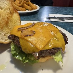 Mushroom Smash Burger - A cheeseburger with cheddar cheese, bacon, lettuce, and onion on a bun, accompanied by onion rings and a burger in the background at a diner.