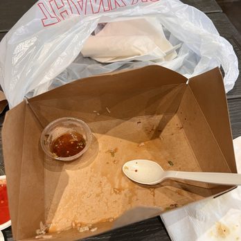 Blue Apsara - A mostly empty brown paper takeout container with some sauce remnants and small food pieces inside. A small clear plastic cup with sauce and a white plastic spoon are also in the container. The container is on a dark table, with a white napkin nearby and a plastic bag in the background. - Cambodian Near Me - Austin, Texas