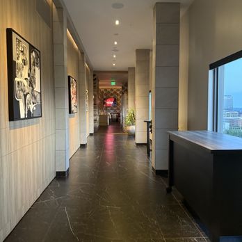 Astra Rooftop - The image depicts a corridor inside a contemporary building, with artwork displayed on the left wall and large windows on the right side allowing natural light to enter. The floor is made of dark, polished material with visible white veining. The ceiling has recessed lighting, and there are decorative plants and furniture at the far end of the hallway, creating a professional and stylish atmosphere. - American Near Me - Columbus, Ohio