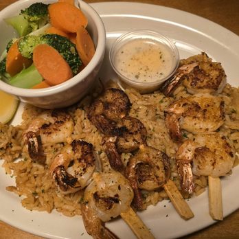 Texas Roadhouse - The image shows a white oval plate with grilled shrimp skewers placed on a bed of rice. The shrimp are seasoned and grilled, with some skewers showing a slight char. A small cup of dipping sauce is positioned near the top of the plate, and a lemon wedge is on the side. A small bowl of steamed broccoli and carrots is also included, placed in a white cup. The plate is on a wooden table, and part of a pen is visible in the top left corner. - Steakhouses Near Me - Eugene, Oregon