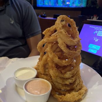 Cheddar's Scratch Kitchen - Onion rings - A tall stack of fried onion rings served with two dipping sauces on a white plate in a restaurant setting. - Comfort Food Near Me - Florence, Kentucky