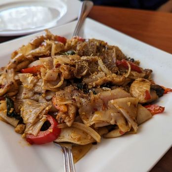 Thai Tip - Spicy noodles with pork - yum! - A square white plate contains stir-fried noodles mixed with vegetables such as red bell peppers, onions, and leafy greens, along with pieces of chicken. The dish appears seasoned with spices and garnished with black pepper. A fork rests on the plate, and a white dish with a spoon is visible in the background, placed on a wooden table. - Thai Near Me - Albuquerque, New Mexico