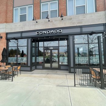 Condado Tacos - Condado restaurant storefront with large glass windows, outdoor seating area with tables and chairs, and a black sign with white lettering. - Tacos Near Me - Carmel, Indiana