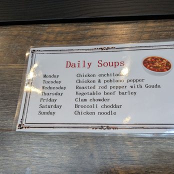 Rooster Run Cafe - Daily soups - A paper menu titled 'Daily Soups' lists different soup options for each day of the week, including chicken enchilada, chicken & poblano pepper, roasted red pepper with Gouda, vegetable beef barley, clam chowder, broccoli cheddar, and chicken noodle. The menu is placed on a wooden surface, and a small, round image of a bowl of soup is located in the top right corner of the menu. - Steakhouses Near Me - Hurricane, Utah