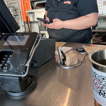 Blaze Pizza - A person in a black uniform is standing at a restaurant counter, holding a smartphone, with a cash register and receipt holder nearby. - Salad Near Me - Ft. Wayne, Indiana