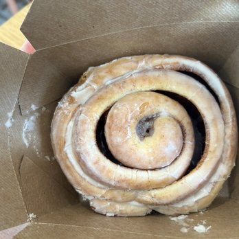 Southpine Bakery - Yummy homemade cinnamon roll to go. - A cinnamon roll with icing is placed inside a brown paper takeout box, showing its spiral shape and some icing on top. - Coffee & Tea Near Me - Eugene, Oregon