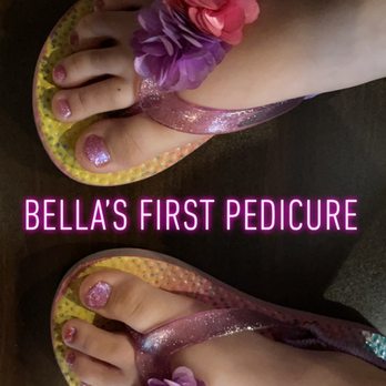 Tracy Nails Spa - Close-up of a child's feet wearing pink and purple floral sandals with glittery toenails. The sandals have large, colorful fabric flowers on the straps. The image includes the text 'Bella's First Pedicure,' suggesting a child's first pedicure experience. The background is dark, highlighting the feet and sandals. - Nail Salons Near Me - Oswego, Illinois