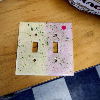 Clay Dreams - Picked up her pottery - A square double light switch cover plate with two rectangular openings is painted with a splatter pattern of green, blue, and pink paint on a white background. It is positioned on a wooden surface, with a basket of newspapers visible in the upper right corner. The setting appears to be a casual indoor environment, possibly a kitchen or workspace. - Paint-Your-Own Pottery Near Me - Arlington, Massachusetts