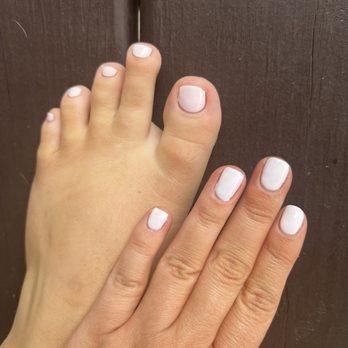 Oasis Nails & Spa - Gel manicure vs regular polish, both #303 - A child's foot with painted toenails is positioned next to an adult's hand with painted fingernails, both with white nail polish, against a wooden background. - Nail Salons Near Me - Bloomington, Illinois