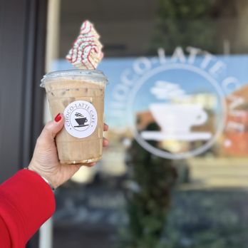 Choco-Latte Cafe - A person with red nail polish and a red sleeve holds an iced coffee in a clear plastic cup with a lid and straw, topped with whipped cream and red syrup. The cup has a Choco-Latte Cafe logo. The background shows a glass window with the café's logo and some blurred outdoor scenery. - Cafes Near Me - Fall River, Massachusetts
