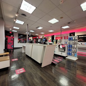 T-Mobile - The photo depicts the interior of a T-Mobile retail store, featuring multiple checkout counters with electronic devices and cash registers. The store has a dark wooden floor, a drop ceiling with fluorescent lighting, and pink signage along the walls indicating sections like phones and accessories. Product displays with phone cases and accessories are visible, and the store appears clean and organized. - Mobile Phones Near Me - Chicago, Illinois