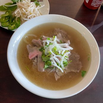 Pholicious - First time here... - A bowl of Vietnamese beef pho with sliced beef, green onions, and cilantro, served in a clear broth. A side plate contains bean sprouts, sliced jalapeños, and lime wedges. - Vietnamese Near Me - Ashburn, Virginia