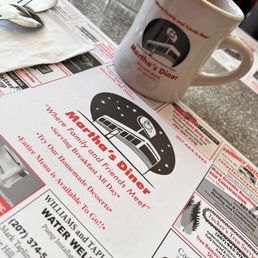 a cup of coffee on a table - A coffee mug with Martha's Diner logo and a menu featuring the diner's name and offerings are on a table at Martha's Diner.