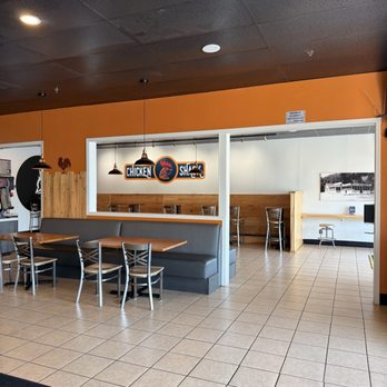 Chicken Shack - Red Bluff - The photo depicts the interior of Chicken Shack, a restaurant with a tiled floor, black ceiling, and orange wall. There is a gray booth with wooden tables and metal chairs in the foreground, and a sign reading 'Chicken Shack' hanging on the wall behind. Additional seating is visible in the background, with a wooden panel and a black-and-white photograph on the wall. - Chicken Shop Near Me - Red Bluff, California