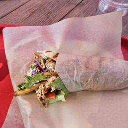 Chicken Gyro - A partially unwrapped chicken gyro with lettuce, tomato, and grilled chicken pieces is placed on a paper-lined tray on a wooden table. A clear plastic cup with a lid and straw, along with a smartphone, are visible in the background. The setting appears to be outdoors or in a casual dining area.