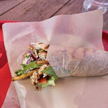 Bentley's Roast Beef - Chicken Gyro - A partially unwrapped chicken gyro with lettuce, tomato, and grilled chicken pieces is placed on a paper-lined tray on a wooden table. A clear plastic cup with a lid and straw, along with a smartphone, are visible in the background. The setting appears to be outdoors or in a casual dining area. - Sandwiches Near Me - Amherst, New Hampshire