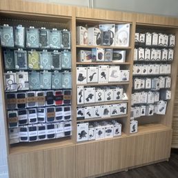 A wooden display shelf holds multiple rows of boxed electronic accessories, including headphones, chargers, and mounts, in a retail store setting.