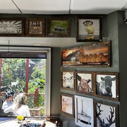 a woman sitting at a table - The image shows an interior wall of Rusty Goat Grill decorated with multiple framed artworks, including animals such as pigs, cows, and a pig with glasses, as well as a barn scene and a giraffe illustration. A window reveals outdoor seating with people and greenery outside. The wall has a greenish paint color, and the artwork is arranged in a gallery style.