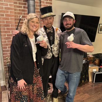 Top Class Magic - Three people are standing inside a room with a brick wall and a television in the background. The person in the middle is dressed as a magician, wearing a top hat, patterned jacket, bow tie, and glittery shoes, and is holding a microphone. The woman on the left has curly blonde hair, a black jacket, and a floral skirt, and is holding a white dove. The man on the right has a beard, a gray t-shirt, a baseball cap, and is also holding a white dove. All three are smiling and appear to be participating in a magic-themed event. - Magicians Near Me